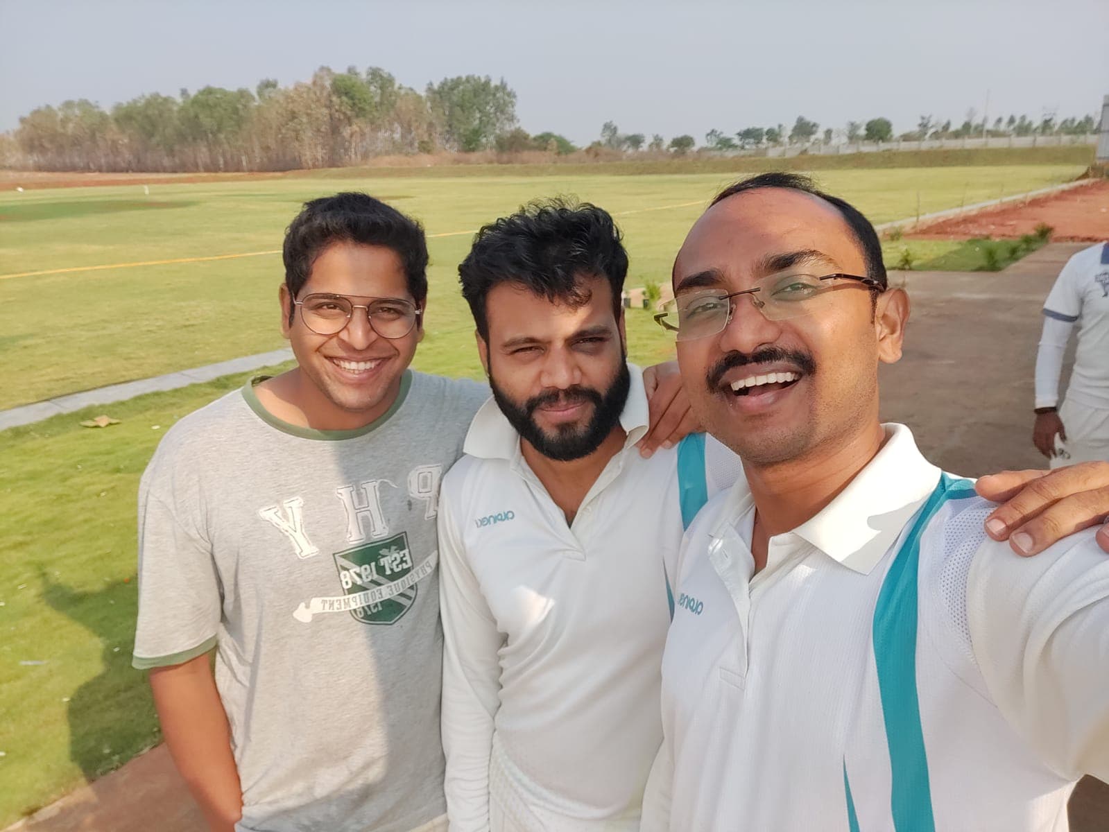 Three Axi Club players in whites taking a selfie on the outfield at Axi Cricket Park